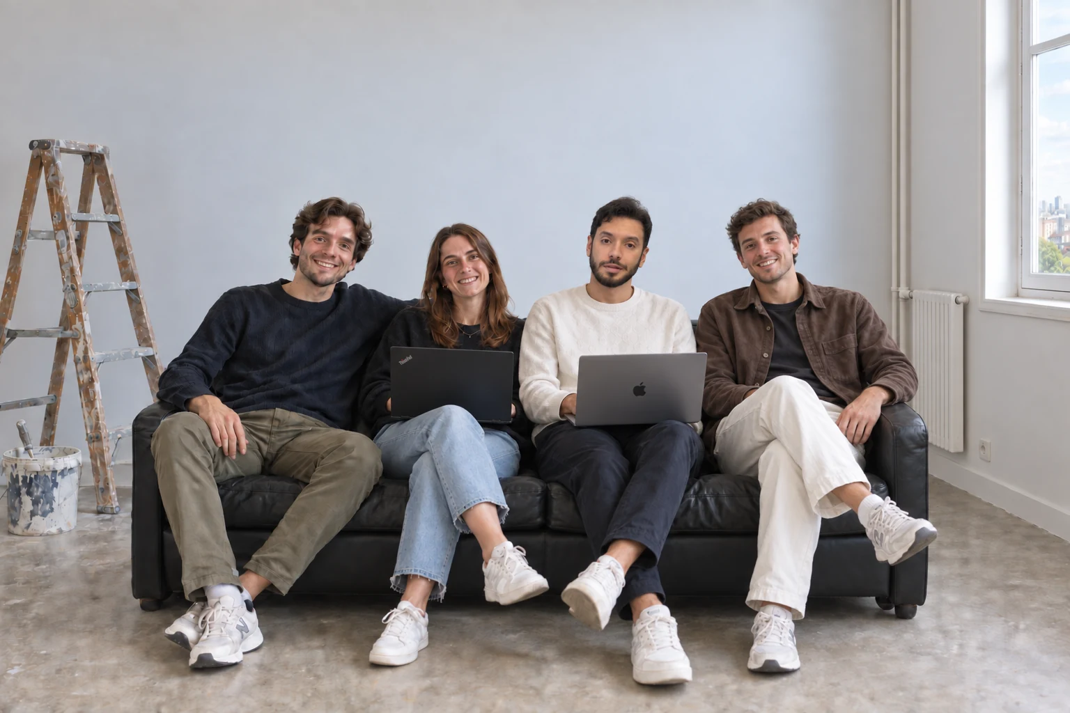 Trois hommes et une femme sur un canapé dans un appartement en rénovation. Deux d’entre eux travaillent sur leur ordinateur portable. Un escabeau et un seau de peinture sont posés à côté.